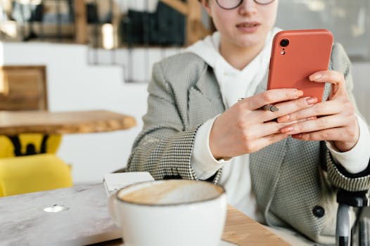 Crop focused female in formal wear and eyeglasses browsing mobile phone while having coffee in cafeteria